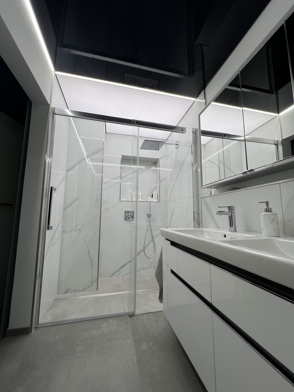 Schwarze Hochglanz-Spanndecke mit umlaufender Lichtlinie über einer Dusche mit Marmorfliesen