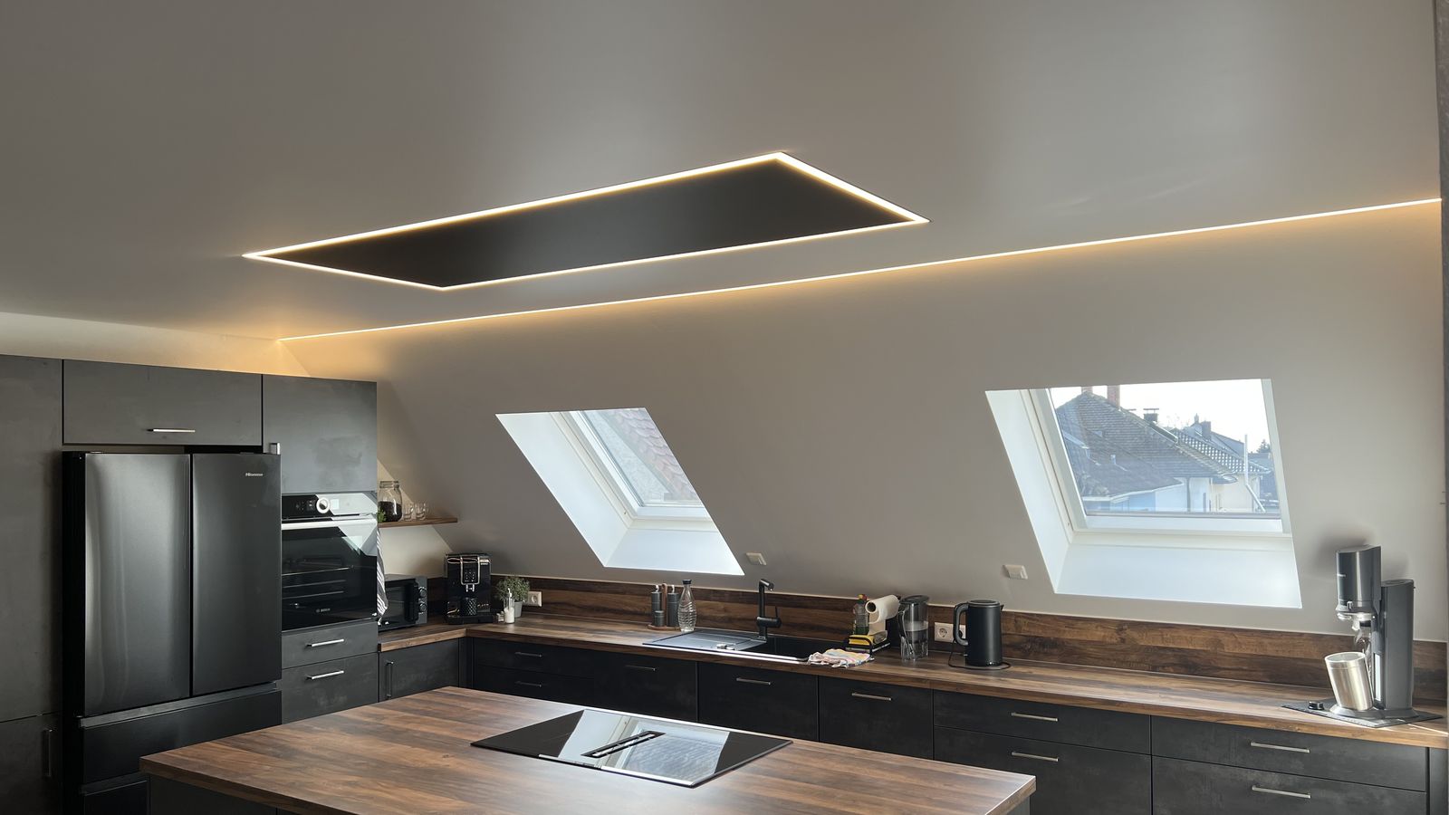 Küche unter der Dachschräge mit matter Spanndecke, schwarzem Lichtfeld über der Kochinsel und Lichtlinie